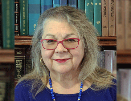 Photograph of presenter Debra Band wearing red glasses, a blue blouse and blue necklace standing in front of a bookcase.