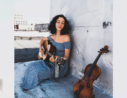 Photograph of Lily Henley seated in front of a marble wall wearing a blue print dress. She plays her guitar with a violin leaning against the wall close by.